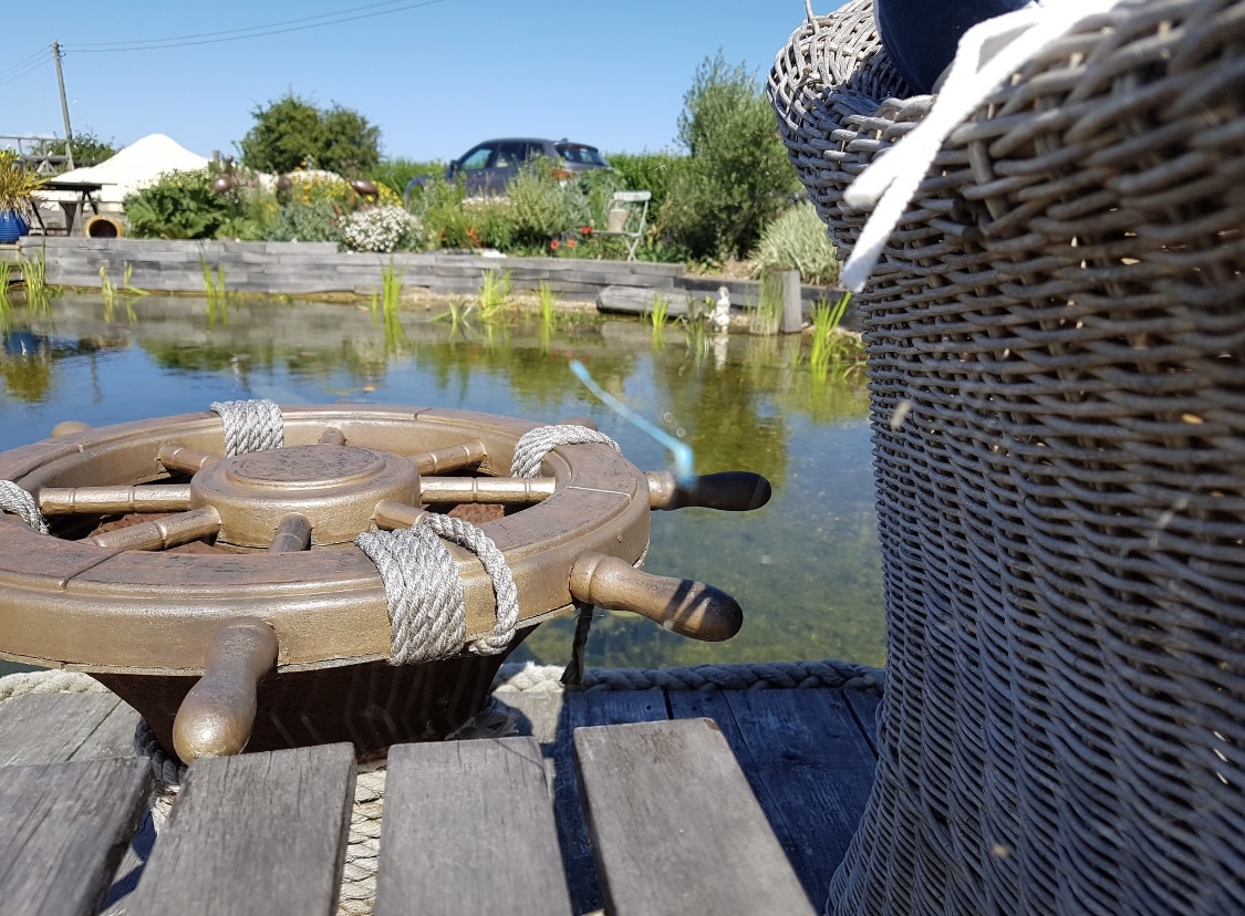 A wooden steering wheel beside a tranquil pond surrounded by greenery.